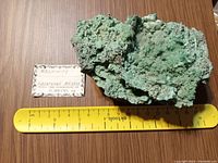 Photo of a green malachite mineral specimen with a yellow ruler beneath for size reference, approximately 7 inches wide. Specimen includes a small label indicating it is from Mexico in excellent condition.