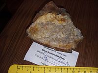 Photo of a small smoky quartz mineral specimen placed on a wooden floor next to a measuring ruler showing about 8 cm width. The specimen is sitting on a small auction bid sheet indicating the species as smoky quartz.