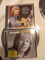 Front view of two sealed John Denver CDs showing cover art. One titled 'Rare and Unreleased Volume 2' with John Denver holding a guitar, the other titled 'The Essential John Denver' with a close-up photo of John Denver.