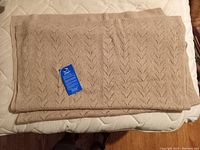 Folded beige scarf with knit chevron pattern and attached blue tag on a mattress.