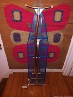 Ironing board folded upright showing blue metal mesh surface and metal handle bar with AC plug connection