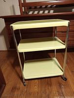 Front side view of the three-tier yellow utility cart with chrome metal frame and caster wheels on a hardwood floor in front of a dresser.
