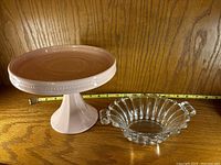 Side-by-side view of the pink ceramic pedestal plate and clear crystal 9-handled bowl with measuring tape showing size.