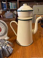 Antique French cream and green enamel Biggin coffee pot with spout and lid shown on wooden table.