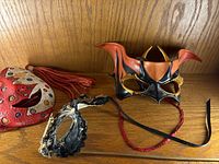 Three masquerade masks displayed on a wooden surface, including the horned leather mask, red and white mask with tassels, and black lace detailed mask.