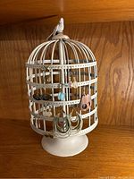 White metal birdcage earring display on wooden shelf with multiple pairs of earrings hung around it. Bird figure on top is visible.