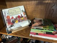 Wood and plexiglass cookbook stand on a wooden shelf with 'The Glass Pantry' cookbook displayed, and five other cookbooks stacked beside it.