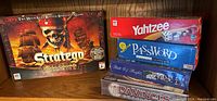 Photo of collection of five boxed family board games including Stratego: Pirates of the Caribbean, Yahtzee, Password, Bill of Rights, and Da Vinci's Challenge stacked on shelf.