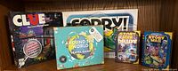 Five family board games including Clue, Sorry!, Around the World Travel Game, Story War, and Story War Deluxe arranged on a shelf.