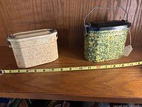 Two antique speckled enamelware lunchboxes side-by-side on wooden shelf with measuring tape showing widths; pink/cream on left and green/yellow on right.