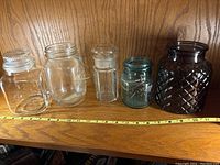 Five glass jars arranged side by side on wooden surface with measuring tape showing size scale.