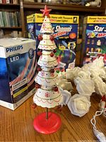 Photo showing vintage tiered metal Christmas tree ornament with red star topper, part of holiday lights lot with Philips LED candy cane bulb pack and two boxed Star Shower Motion laser light projectors in background, plus string of white paper rose lights visible.
