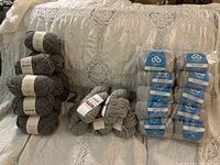 Lot of multiple yarn skeins packaged and loose on surface, showing quantity and general grey tones.