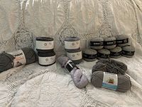 Full view of lot with all skeins displayed on fabric surface showing their labels and colors varying in shades of grey and one lavender/silver skein