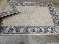Two flat outdoor rugs laid on floor showing blue floral border on tan background.