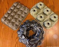 Three NordicWare pans on a wooden table: Floral petite four pan, petite bundt pan, and decorative wreath bundt pan