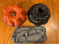Orange Nordic Ware Fiesta Bundt pan, black Nordic Ware Chrysanthemum Bundt pan, and John Wright cast iron sea life cookie mold on wood table.