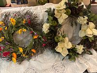 Two wreaths displayed on a white cloth background - one with white flowers and pine cones, the other with colorful flowers on twigs
