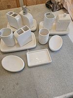Overview of all 13 white ceramic bathroom accessory items arranged on a surface, two trays with multiple holders, soap dishes, and a dispenser included.