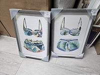 Two bikini prints framed with silver frames, positioned upright on the floor, showing tropical leaf patterned bikini tops and bottoms.