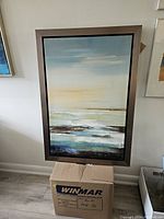 Framed ocean print leaning against wall atop a cardboard box, showing the full artwork and frame