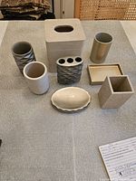 Photo showing 9 of the 12 bathroom accessory pieces including tissue box cover, toothbrush holder, soap dish, and various containers with neutral and textured finishes.