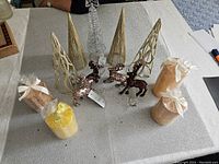 Photo of five gold glitter cone-shaped Christmas tree decorations, four small reindeer figurines in glittery bronze and gold, and four wrapped candle gift boxes tied with ribbon bows on a white tablecloth.