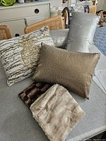 Four decorative throw cushions, including beige and gold patterned square, gold textured lumbar, and two faux fur cushion covers in brown and beige.