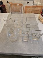 Eight clear glass vases displayed on table in various shapes including cylindrical, square, and rectangular forms.