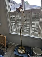 Full view of the 54 inch adjustable brass floor lamp standing upright near window with white shutters. Shows adjustable arm and lampshade with brass finish.