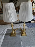 Pair of polished brass candlestick lamps with white pleated shades shown on a table with background furniture.