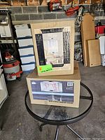 Two Metz light fixtures in original boxes on a circular glass table indoors in a garage/utility room setting, a vacuum and boxes in background.