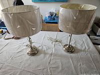 Two silver metal table lamps with white drum shades displayed on a table, showing overall condition and appearance.