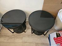 Pair of two round end tables with black glass tops, black metal frames, and wood lower shelves with marbled design placed side by side on a laminate floor.