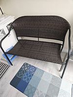 Front view of a metal and vinyl 2-seater outdoor loveseat showing woven seating and black metal frame on a patterned indoor floor.