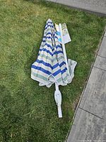 Folded patio umbrella with a striped canopy in white, blue, green, and beige stripes lying on grass beside a stone pathway.