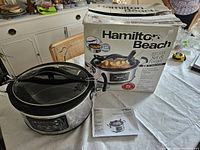 Photo of Hamilton Beach slow cooker unit with glass lid, partially closed, next to original box and instruction manual on table.