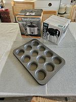 Full set showing Toastmaster slow cooker box, Oggi thermal coffee carafe box, and metal 12-cup muffin tin on a table.
