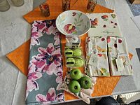Display of spring-themed table linens, placemats, napkins, salad bowl, battery lights shaped like green apples, apple wax candles, and fabric trivet on a table.