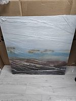 Front view of the square water and landscape canvas print wrapped in plastic, showing abstract blue, brown, and white tones.