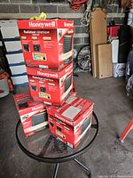 Five red Honeywell ceramic heater boxes stacked on glass round table in garage, assorted background storage items.