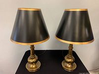Pair of brass table lamps shown on a black surface with black wall background, clear view of shape and color.