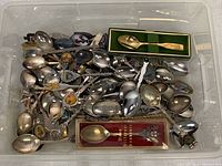 Wide view of mixed assorted collector spoons piled in a container, showing multiple metal spoons with decorative handles and crests, including boxed spoons.