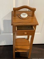 Front view of an antique blonde wood small table with a handle on top, open drawer beneath, and a lower shelf. The table features a ceramic inkwell set into a circular wooden holder on the top surface.