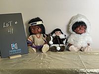 Front view of all three dolls seated side-by-side on a beige surface with a black lot number sign on left