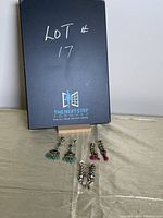 Photo showing three pairs of drop earrings laid out on fabric beneath a black box with writing Lot #17.
