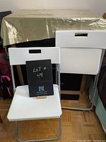 Two white IKEA Gunde folding chairs, one fully unfolded and the other folding against a covered table and wall.