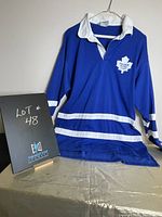 Blue men's rugby shirt with white collar and stripes, Toronto Blue Leafs maple leaf logo on chest, hung on hanger with chalkboard showing lot number.