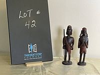 Two tall carved wooden Masai statues, male and female, standing upright with a seller lot board behind.