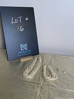 Set of multi-strand faux pearl necklace, matching bracelet, and stud earrings on beige cloth with 'Lot 16' sign in background